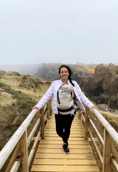 A young woman hiking through Fuerteventura, Spain while holding her baby in a baby carrier while crossing a narrow bridge.
