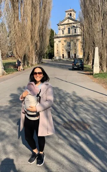 A young woman standing in front of a yellow castle in Varese, Italy. The woman is holding the baby in the front with a baby carrier.