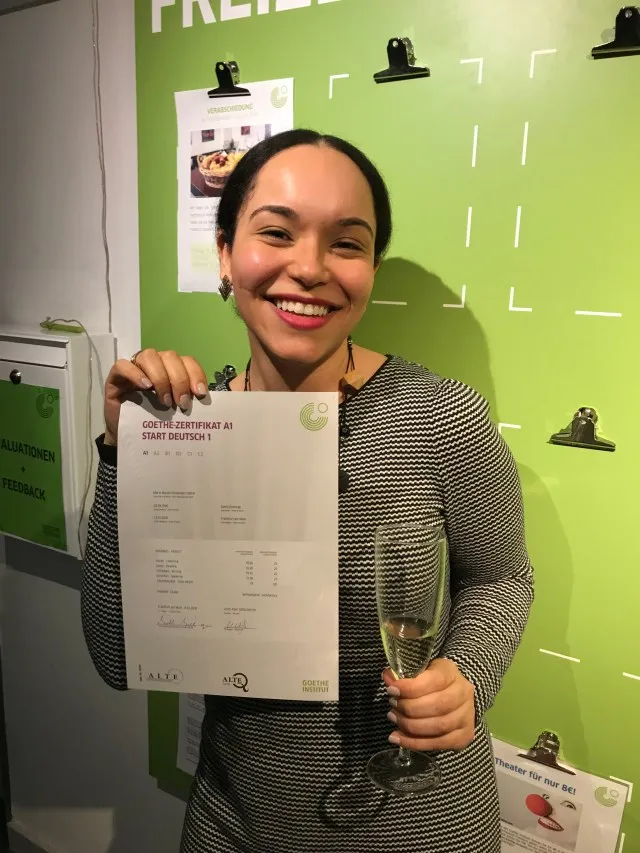 A young woman in her 30's proudly holding a language certificate from Goethe Institute. Celebrating with a glass of champagne achieving her A1 German language certificate.