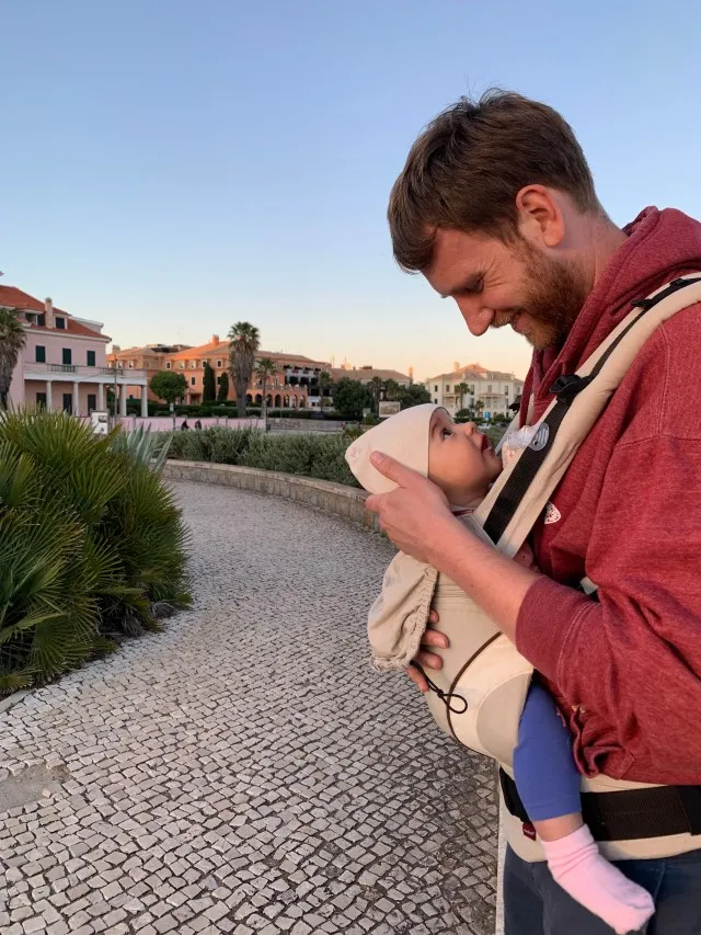 Dad holding his small daughter in a baby carrier while they look up at each other.