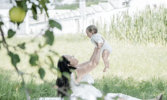 Mother holding child in the air above her. Both mother and daughter wearing white while sitting on the grass. Green scenery and nature surrounds them.